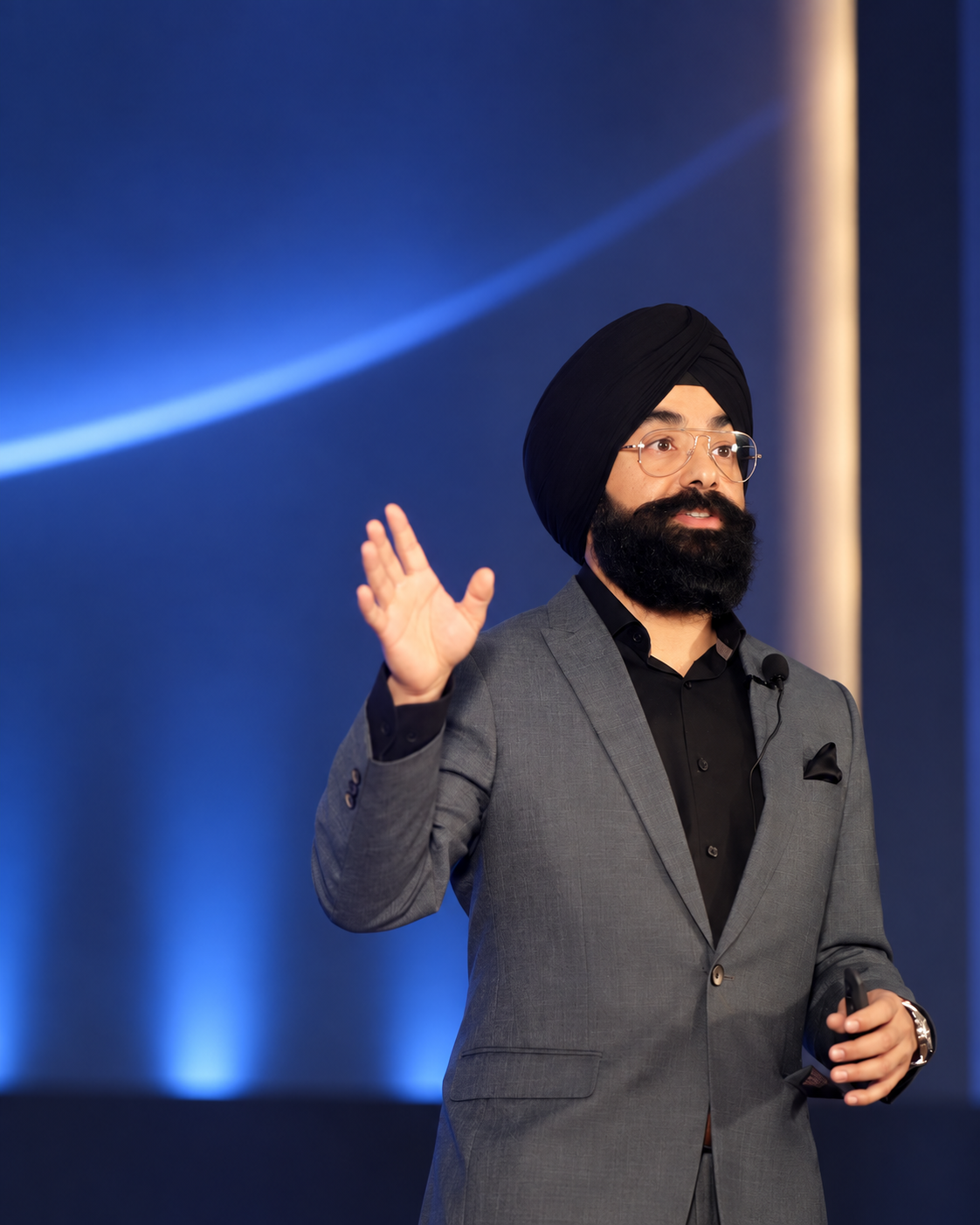Navdeep Chhabra speaking on a blue-lit stage in a grey blazer.